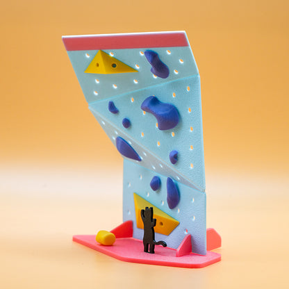 Non Vertical Lab rock climbing toy and gift – A black cat figurine gazes at a vibrant mini climbing wall with blue holds and yellow volumes. A perfect collectible for climbing enthusiasts and a fun gift for all ages.